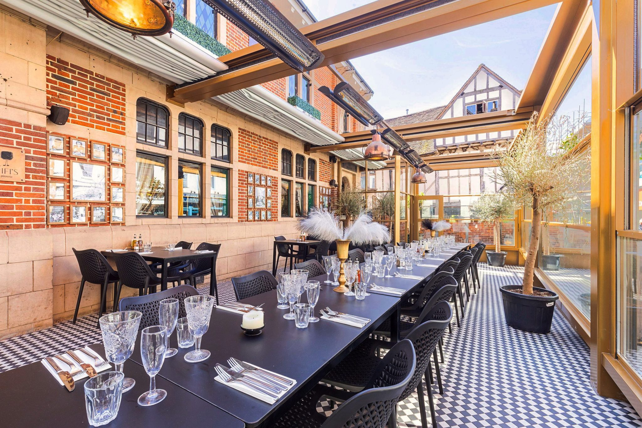 Bright and elegant party venue with long black tables, crystal glassware, decorative centerpieces, checkered flooring, and glass walls offering natural light and charming views.